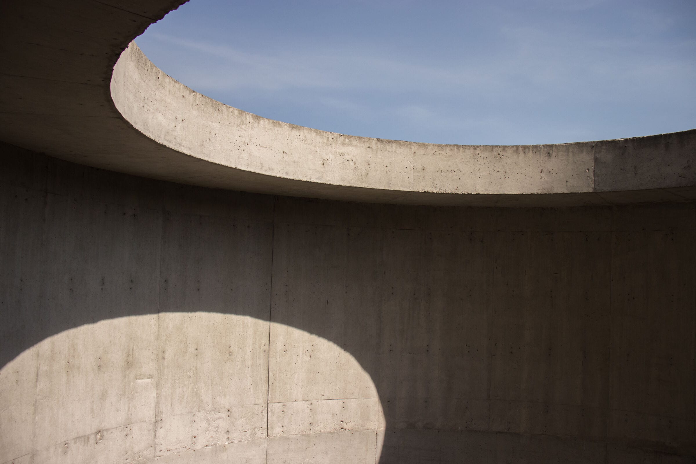 A curved board-marked concrete wall opening to clear sky, with a soft circular shadow cast across it.