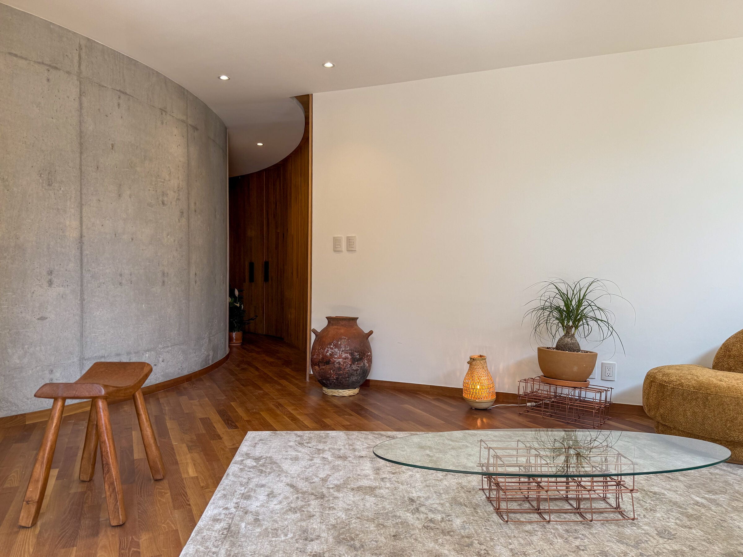 A living room where a curved concrete wall meets a white plaster wall, with a wooden stool, clay vessel and round glass table on a pale rug.